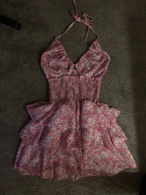 Blommig rosa kortklänning med volanger - Supersöt kortklänning med blommigt mönster i rosa och vita toner. Klänningen har omlott-topp med knyt i nacken, smockad midja och flera lager volanger i kjolen. Perfekt för sommarens alla tillfällen och ger en romantisk vibe.