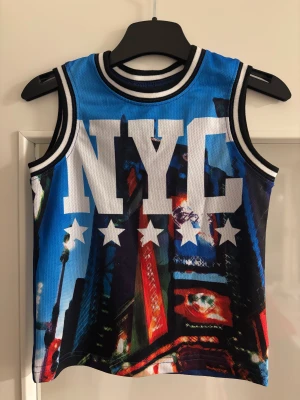 Blå NYC linnetopp med tryck - Säljer en cool blå linnetopp med NYC-tryck och stjärnor framtill. Toppen har ett färgstarkt motiv av stadsmiljö och kontrasterande svarta och vita ränder vid hals och ärmhål. Perfekt för dig som gillar streetstyle och sportig vibe.