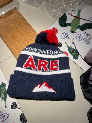 Mörkblå Åre mössa med tofs - Snygg mörkblå stickad Åre-mössa med vit och röd broderad text,  Mössan har en stor tofs i mörkblått och rött på toppen och vita ränder runt om. Perfekt för kalla dagar.