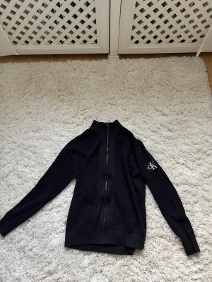 Svart stickad kofta från Calvin Klein - Svart stickad kofta från Calvin Klein Jeans med dragkedja framtill och ribbad krage. Snygg CK-logga på vänster ärm. Köpt för ny pris 1400 kr, aldrig använt. XS/S