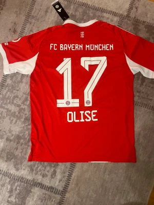 Bayern München matchtröja Olise #17 - Säljer en officiell FC Bayern München fotbollströja från Adidas, med Olise och nummer 17 på ryggen. Tröjan är röd med vita detaljer, har korta ärmar och ett snyggt randigt mönster framtill. Materialet är lätt polyester med Aeroready-teknologi för bästa komfort.