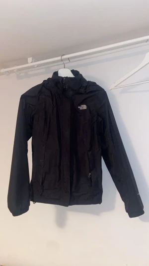 Svart vindjacka från The North Face - Svart vindjacka från The North Face med huva, dragkedja och kardborreknäppning framtill. Jackan har två sidofickor med dragkedja och är tillverkad i lätt polyester. Enda problemet med jackan är att ena fram-fickan inte går att stänga och en kardborre lossnat lite. Jag fixn jackan av min familj så jag har inget som intygar på att den är äkta!