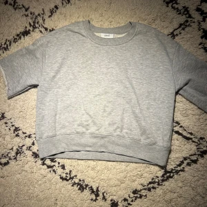 Grå croppad sweatshirt  - Grå croppad sweatshirt från Crunt med korta ärmar och rund hals. Tröjan har en loose passform och är tillverkad i mjuk bomull. Perfekt för chill dagar och enkel att matcha med jeans eller joggers.