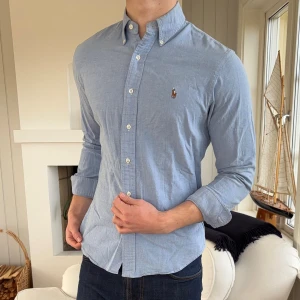 Blå slim fit skjorta från Ralph Lauren - Snygg ljusblå skjorta från Ralph Lauren i slim fit-modell, tillverkad i stretchig bomull. Klassisk button-down krage och broderad logga på bröstet. Långärmad med vita knappar framtill och vid ärmslut. Perfekt för dig som gillar stilrena och tidlösa plagg.