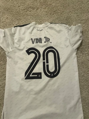 Real Madrid Vini Jr. 20 matchtröja - Säljer en officiell Real Madrid fotbollströja från Adidas med Vini Jr. och nummer 20 på ryggen. Tröjan är vit med svarta detaljer, klubbmärke på bröstet och sponsortryck 'Emirates Fly Better'. Tillverkad i lätt och ventilerande material, perfekt för träning eller match. Gammal storlek xs