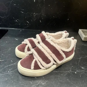 Bruna/ vinröda sneakers från Inuikii med teddy - Unika sneakers från Inuikii i mörkbrunt mocka med fluffiga beige teddy-detaljer. Snygg design med breda kardborreband och vit platt sula. Insidan är fodrad med mjukt teddy-material för extra komfort. Perfekt för dig som gillar statement-skor med cozy vibe.