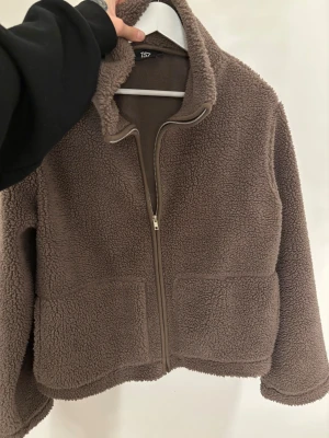 Brun teddyjacka med dragkedja - Mysig brun teddyjacka från 157 med hög krage och hel dragkedja framtill. Jackan har två stora fickor och är tillverkad i fluffigt fleece-material som ger en chill och avslappnad vibe. Perfekt för kalla dagar när du vill vara både varm och snygg. St M men sitter bra på mig som är s-xs