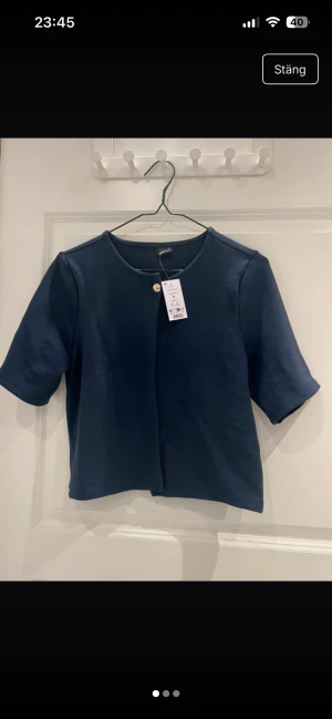 Mörkblå kofta från Gina Tricot - Snygg mörkblå kofta från Gina Tricot med korta ärmar och rund hals. Koftan har en enkel design med en guldfärgad knapp upptill och är tillverkad i mjuk bomull. Perfekt för dig som gillar stilrena plagg med en twist.