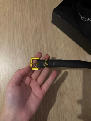 Svart skärp från Saint Laurent - Säljer ett stilrent svart bälte från Saint Laurent med guldspänne och YSL-logga i metall.