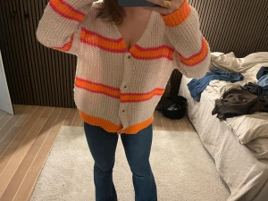 Randig kofta med orange och rosa detaljer - Säljer en loose-fit stickad kofta med breda ränder i orange och rosa på en beige bas. Koftan har v-ringning, knappar framtill och långärmade ärmar med orange muddar. Perfekt för dig som gillar färgglada och chill plagg.