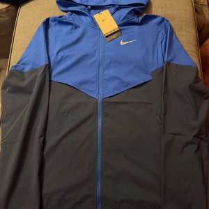Nike blå och svart vindjacka M - Nike vindjacka i blått och svart med huva och hel dragkedja framtill. Jackan har meshfoder, praktisk innerficka med dragkedja och snyggt Nike Swoosh på bröstet. Perfekt för löpning och träning, lätt och sportig design.
