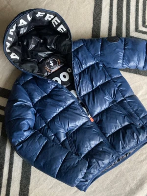 Pufferjacka från Save The Duck - Säljer en blå pufferjacka från Save The Duck med huva och coolt tryck på insidan. Jackan har två fickor med dragkedja samt Save The Duck-logga på ärmen. Perfekt för dig som vill ha en lätt och snygg jacka med street vibe. Ps ytterst liten defekt på dragkedjan vid ansiktet men påverkar inte funktionen(syns på sista bilden)