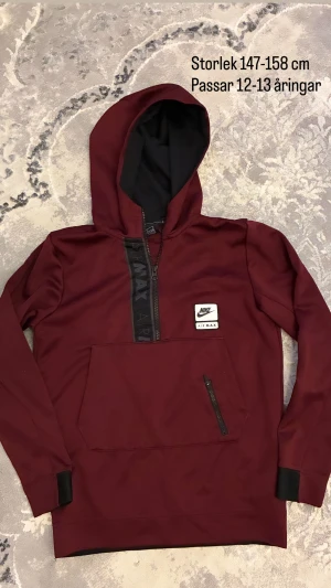 Vinröd Nike Air Max hoodie med half zip - Snygg vinröd hoodie från Nike Air Max med svart half zip och stor ficka framtill med dragkedja. Tröjan har huva, långa ärmar och svart detalj vid muddar. Perfekt för dig som gillar sportig stil och vill ha något bekvämt och trendigt.