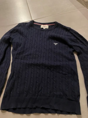 Mörkblå kabelstickad tröja Hampton Republic - Snygg mörkblå kabelstickad tröja från Hampton Republic med rund hals och diskret logga broderad på bröstet. Tröjan har långärmad passform och är gjord i mjuk bomull, perfekt för dig som gillar klassisk stil med en modern twist.