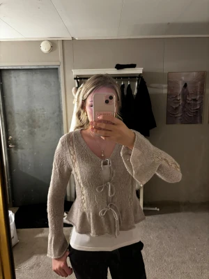 Beige stickad kofta med knyt - Supersöt beige stickad kofta med v-ringning och tre knyt framtill. Koftan har vida ärmar, spetsdetaljer längs sidorna och ett fint hålmönster både fram och bak. Perfekt för lager-på-lager och ger en romantisk vibe.