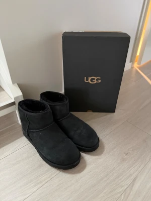 Svarta UGG boots i mocka - Säljer ett par klassiska svarta UGG boots med rund tå och platt sula. Bootsens utsida är i mjuk mocka och insidan är fodrad med fluffig päls för extra värme. Perfekta för kalla dagar och har UGG-logga baktill. Snygg och enkel design som passar till allt.