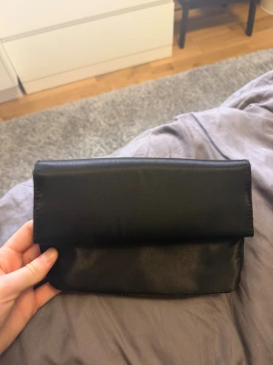 Svart clutchväska - Stilren svart kuvertväska med magnetlås. Väskan har två fack på insidan och en enkel, rektangulär form. 