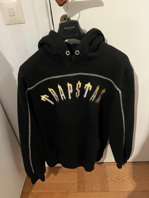 Svart Trapstar hoodie med gult tryck - Svart hoodie från Trapstar i storlek M med metallicfärgat tryck i guld och silver över bröstet. Tröjan har huva med snörning, vita kontrastsömmar längs axlar och ärmar samt en stor magficka. Perfekt för dig som gillar streetwear och vill sticka ut. Hör av dig vid minsta fundering! Pris kan diskuteras vid snabb affär 