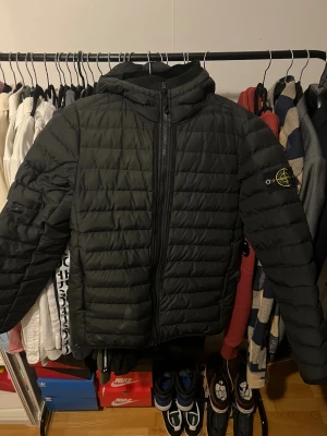 Svart dunjacka från Stone Island - Svart dunjacka från Stone Island med huva och klassisk patch på vänster arm. Jackan har en lagning på höger arm och en rivning på väster ficka men är i gott skick annars.