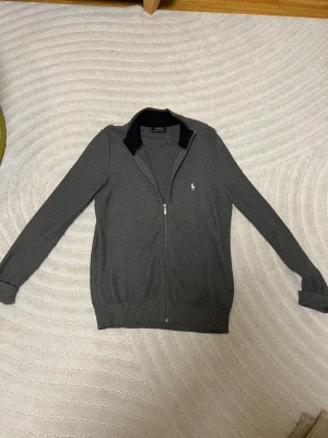 Grå kofta från Polo Ralph Lauren - Snygg grå kofta från Polo Ralph Lauren med dragkedja och ribbad svart krage. Klassisk broderad logga i vitt på bröstet. Långärmad och tillverkad i mjuk bomull, perfekt för lager-på-lager. Passar dig som gillar stilrena och tidlösa plagg.