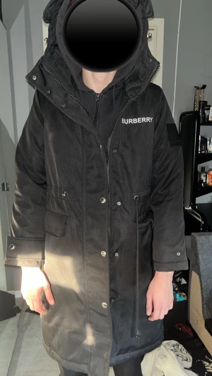 Svart Burberry parkajacka M - Parkajacka från Burberry i svart med logga på bröstet. Jackan har huva, snörning i midjan, tryckknappar och dragkedja. Insidan är quiltad med klassiskt Burberry-rutigt mönster i beige, rött, svart och vitt. Materialet är bomull och polyester, perfekt för kyliga dagar.