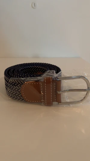 Mörkblå randigt flätat bälte - Snyggt mörkblått flätat bälte med vita ränder och detaljer i brunt skinn. Spännet är i silverfärgad metall och bältet har en cool, modern look som passar till jeans eller chinos. Perfekt accessoar för att lyfta din outfit med lite extra stil.