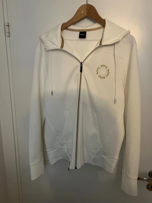 Vit hoodie från BOSS med dragkedja - Vit hoodie från BOSS med gulddetaljer och logga på bröstet. Tröjan har huva med snörning, hel dragkedja och ribbade muddar. Materialet är mjukt och känns som bomull, perfekt för chill dagar. Snyggt guldtryck på insidan av kragen och stilren design. Aldrig använt, inget tecken på användning, nyttpris 3500-4000 mitt 1100