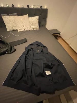 Mörkblå väst med huva från Lyle & Scott - Snygg mörkblå väst från Lyle & Scott med huva och stilren design. Västen har en enkel, rak passform och är tillverkad i mjukt material som känns skönt mot kroppen. Perfekt för dig som gillar en clean look och vill ha något som funkar till många outfits. Kejdan e trasig 