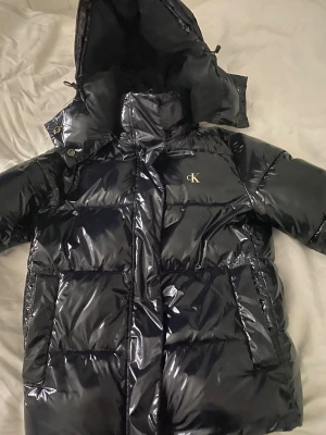 Svart pufferjacka från Calvin Klein - Säljer en svart, glansig pufferjacka från Calvin Klein med huva och dragkedja. Jackan har CK-logga på bröstet, stora fickor och quiltad design. Perfekt för kalla dagar med sin rejäla vaddering och snygga look. Nästan helt ny och andvänd fåtal gånger