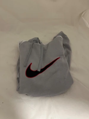 Grå Nike hoodie med svart Swoosh - Snygg grå hoodie från Nike med stor svart Swoosh-logga med röd kant på bröstet. Tröjan har huva med snörning och långa ärmar, samt en klassisk känguruficka framtill. Perfekt för chill eller streetwear-stil.