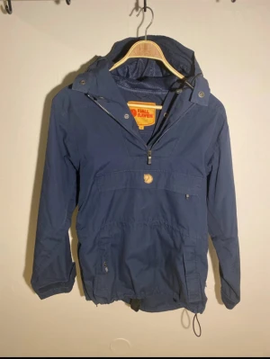 Blå Anorak från Fjällräven  - Säljer en blå vindjacka från Fjällräven i storlek XS. Den är stor i storleken! Tycker mer den sitter som en M om man inte har mellanlager under. Jackan har huva, halv dragkedja framtill, flera fickor och justerbar nederkant. Tillverkad i en mix av polyester och bomull, perfekt för dig som vill ha en snygg och funktionell jacka till vardags.
