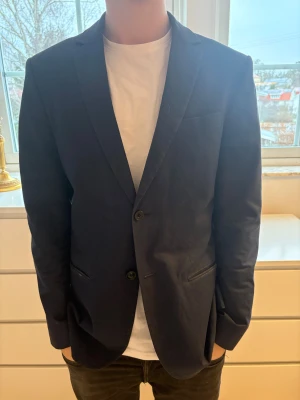 Mörkblå kavaj från Filippa K - Snygg mörkblå kavaj från Filippa K i klassisk modell med två knappar framtill och smala slag. Kavajen har diskreta fickor och en stilren passform som ger en clean look. Perfekt för dig som vill ha en enkel men stilfull outfit.