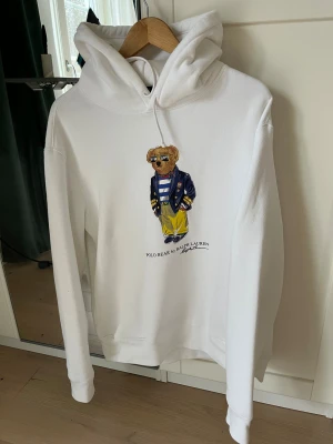 Vit hoodie Polo Bear Ralph Lauren L - Vit hoodie från Polo Ralph Lauren med ikoniskt Polo Bear-tryck på bröstet. Tröjan har huva med snörning och långa ärmar. Materialet är mjukt och känns som bomull, perfekt för chill dagar. Klassisk loose passform och snyggt tryck som sticker ut.