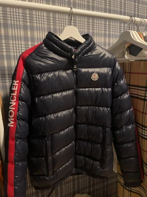 Moncler mörkblå pufferjacka - Säljer en mörkblå pufferjacka från Moncler med röd insida och bred röd rand längs ärmen där Moncler-loggan är broderad i vitt. Jackan har dragkedja framtill, ståkrage och två fickor med knappstängning. Perfekt för kalla dagar.