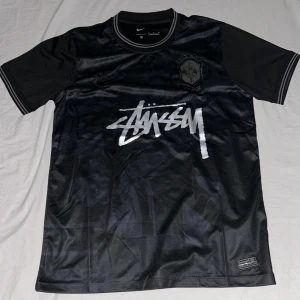 Svart Brasil Nike x Stüssy fotbollströja/jersey - Snygg svart Brasil fotbollströja från Nike x Stüssy med silvrigt Stüssy-tryck på bröstet och broderat Brasil-märke. Tillverkad i Dri-FIT material som andas, med rund hals och korta ärmar. Subtila mönster i tyget och ribbade detaljer vid hals och ärmslut.