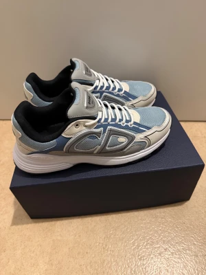 Dior B30 sneakers i blått och vitt - Säljer ett par Dior B30 sneakers i en snygg mix av ljusblått, vitt och grått. Skorna har mesh och skinnpaneler, vita snören och en chunky vit sula. CD-logga på hälen och Dior-märke på plösen. Perfekt för dig som gillar exklusiva och trendiga sneakers.