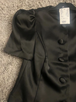 Svart blus med puffärm och stora knappar - Säljer en svart blus från H&M i glansigt satinmaterial. Blusen har korta puffärmar och stora tygklädda knappar framtill. Figurnära passform med markerad midja och rund halsringning. Perfekt för dig som gillar stilrena och trendiga detaljer.