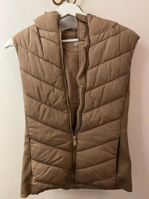 Beige quiltad dunväst från Zara - Snygg beige dunväst från Zara med quiltad design och huva. Västen har dragkedja framtill och ribbade sidor för extra komfort. Perfekt att slänga över en hoodie när det är kyligt ute. Stilren look som funkar till många outfits.