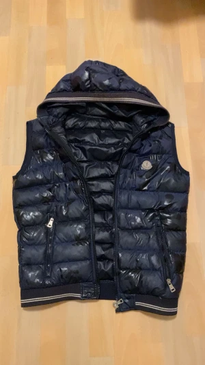Mörkblå dunväst från Moncler - Snygg mörkblå dunväst från Moncler med huva och quiltad design. Västen har två dragkedjefickor fram, ribbad kant med vita ränder och Moncler-logga på bröstet. Perfekt för lager-på-lager och streetstyle. Fylld med dun och har glansig finish.