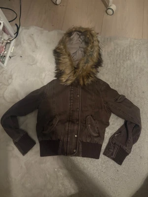 Brun bomberjacka med fuskpäls från Gabrielle's Vault - Snygg brun bomberjacka från Gabrielle's Vault med fluffig fuskpäls på huvan och mjukt beige foder. Jackan har ribbade muddar, dragkedja och knappar framtill samt praktiska fickor med lock. Perfekt för dig som gillar streetstyle och vill ha en varm jacka med coola detaljer.