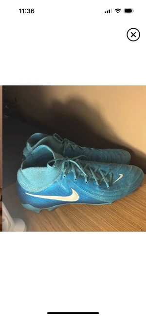 Nike blå fotbollsskor med snörning - Säljer ett par blå Nike fotbollsskor med vit swoosh på sidan. Skorna har snörning och är tillverkade i syntetmaterial med stickad flyknit-krage för extra komfort. Sulan har dobbar för bra grepp på planen. Perfekt för dig som vill ha snygga och funktionella skor till matchen. Storlek 44