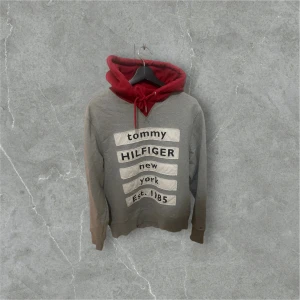Grå och röd hoodie Tommy Hilfiger M - Snygg grå hoodie från Tommy Hilfiger med röd huva och snörning. Framsidan har stora vita tryck med texten 'tommy HILFIGER new york Est. 1985'
