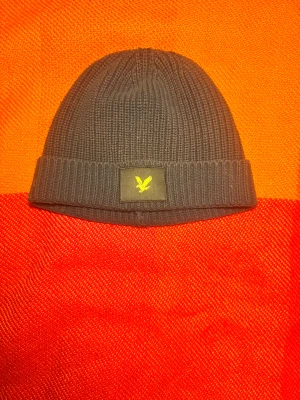 Mörk blå ribbad mössa från Lyle & Scott - Mörk blå stickad mössa från Lyle & Scott med ribbad design och uppvikt kant. Märkets klassiska gula fågel-logga framtill på en svart patch. Perfekt för kalla dagar och enkel att matcha med din jacka.