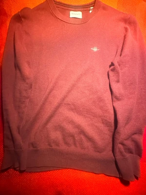 Bordeaux stickad tröja från GANT - Snygg bordeauxfärgad stickad tröja från GANT med rund hals och diskret logga broderad på bröstet. Tröjan har långärmad passform och ribbade muddar vid ärmslut och nederkant. Perfekt för dig som gillar klassisk stil med en modern touch.