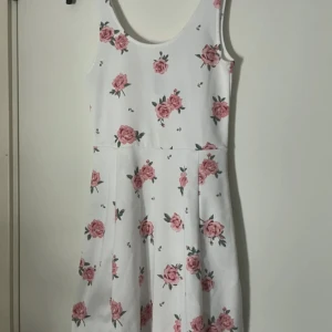 Vit blommig sommarklänning - Superfin vit klänning med rosa blommor och ärmlös design. Klänningen har en rundad halsringning och ett lätt utställt kjolparti som ger en söt och feminin look. Perfekt för varma dagar och somriga vibes.