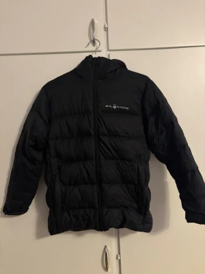 Svart pufferjacka från Sail Racing - Svart pufferjacka från Sail Racing med huva och dragkedja framtill. Jackan har quiltad design och logga på bröstet samt en detalj på huvan. Perfekt för kalla dagar och riktigt snygg till streetwear-stilen.