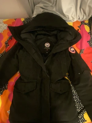 Svart dunjacka från Canada Goose - Säljer en svart dunjacka från Canada Goose med huva och snygg pälskant. Jackan har dragkedja, stora fickor framtill och klassisk patch på ärmen. Perfekt för kalla dagar och riktigt stilren look. PRIS KAN DISKUTERAS!!!!