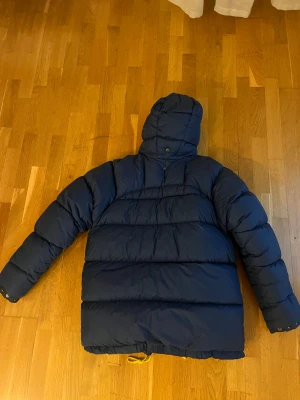 Blå dunjacka från Fjällräven med huva - Säljer en blå dunjacka från Fjällräven med huva, gul snörning och dragkedja framtill. Jackan har två stora fickor med knappar och Fjällräven-logga på bröstet. Perfekt för kalla dagar och har en riktigt schysst oversized look.
