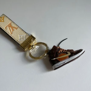  Vuitton sneaker nyckelring - Exklusiv Vuitton nyckelring med beige och guldigt band i präglat material och en miniatyr av en brun sneaker med klassiskt -mönster. Detaljer i vitt, orange och guld samt guldfärgad ring. Perfekt accessoar för dig som älskar streetstyle och lyx.