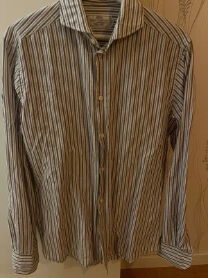 Randig linneskjorta från These Glory Days - Snygg skjorta med bruna, vita och blå ränder från These Glory Days. Modellen är med klassisk krage och knappar framtill. Långärmad och tillverkad i mjuk bomull och linne, perfekt för dig som gillar stilren design med skandinavisk touch.Passar mig bra för er som är 175-185